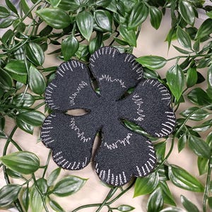Könnte beinhalten: Schwarze Stoffblume mit weißen Nähten, die einem vierblättrigen Kleeblatt ähnelt. Die Blume ist flach und hat eine leicht strukturierte Oberfläche.