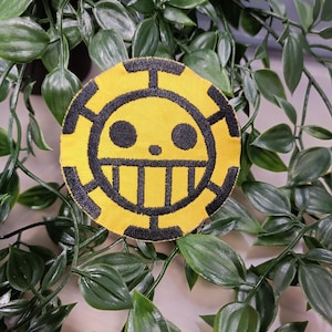 Könnte beinhalten: Ein gestickter Patch in Gelb und Schwarz mit einem Totenkopf- und Knochenkreuz-Motiv. Der Patch ist rund und hat einen schwarzen Rand.