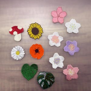 Pode incluir: Uma coleção de patches bordados com vários desenhos florais e botânicos. Inclui um cogumelo vermelho e branco, um girassol amarelo, uma margarida branca, uma papoila laranja, folhas verdes e patches de flores em rosa, branco e roxo.