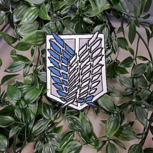 May include: A white and grey embroidered patch with blue accents depicting a pair of wings. The wings are stylised and have a geometric design. The patch is surrounded by green leaves.