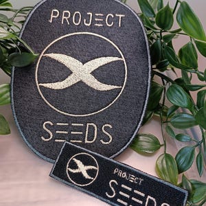 Könnte beinhalten: Zwei schwarze gestickte Patches mit weißen Nähten. Der größere Patch ist oval geformt und sagt "PROJECT SEEDS" mit einem stilisierten "X" in der Mitte. Der kleinere Patch ist rechteckig und sagt "PROJECT SEEDS" mit einem stilisierten "X" in der Mitte.