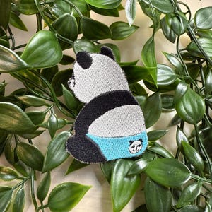 Könnte beinhalten: Ein schwarz-weißes Pandabärchen, das blaue und weiße Unterwäsche trägt. Der Panda sitzt auf seinen Hinterbeinen mit dem Rücken zum Betrachter. Der Panda hat ein kleines Pandagesicht auf seinem Hinterteil.