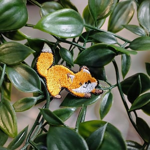 May include: A small embroidered patch featuring a cartoon fox with a white belly and orange fur. The fox is curled up with its tail behind it. The patch is set against a green leafy background.