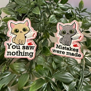 Könnte beinhalten: Zwei gestickte Aufnäher mit Cartoon-Katzen mit Messern. Einer ist hellbraun mit dem Text "You saw nothing", der andere ist grau mit "Mistakes were made". Beide haben rosa Ränder und rote Blutdetails.