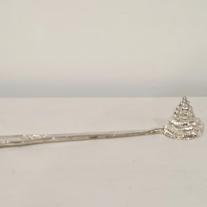 Puede incluir: Una cuchara de metal plateado con un mango largo y delgado y un cuenco en forma de estructura cónica escalonada con crestas concéntricas. La superficie metálica tiene un acabado reflectante y el diseño es consistente desde el mango hasta la punta.