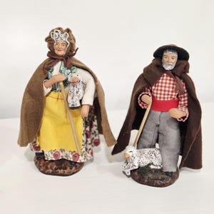 May include: Two handcrafted figurines of a shepherd and shepherdess. The shepherdess wears a yellow skirt, floral apron, and brown cape. The shepherd holds a staff and a lamb figurine. The shepherd wears a red and white checkered shirt, gray pants, and a brown cape.