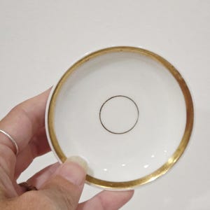 May include: A small, round white ceramic saucer featuring a wide, metallic gold-colored rim along the outer edge and a single, thin, dark-colored circular line centered in the middle of the smooth, white interior surface of the dish.