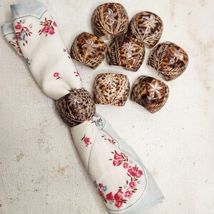 Könnte beinhalten: Eine Sammlung dekorativer Serviettenringe und eine Serviette mit floralem Muster. Die Serviettenringe sind braun mit einem gesprenkelten Muster und geschnitzten floralen Designs. Die Serviette ist weiß mit rosa und blauen floralen Akzenten.