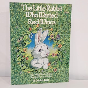 Puede incluir: Un libro infantil titulado "The Little Rabbit Who Wanted Red Wings". La portada presenta un conejo blanco rodeado de una corona floral sobre un cielo azul, en un fondo de hierba verde. Es un libro Cricket.