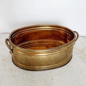 May include: An oval-shaped brass planter with two handles. The planter is empty and has a shiny, polished finish.