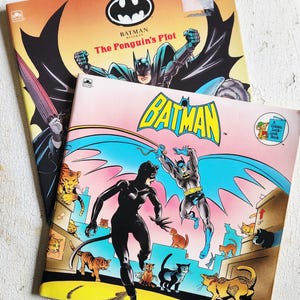 May include: Two vintage Batman comic books. The top book is titled "The Penguin's Plot" with Batman in a dynamic pose. The bottom book, "The Purrfect Crime," features Catwoman and Batman with a pink and yellow background.