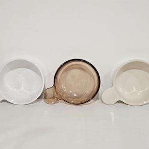 Könnte beinhalten: Drei kleine, runde Schalen mit angebrachten Griffen. Zwei sind weiß und eine ist transparent bernsteinfarben mit einem dunkelbraunen Rand. Die Schalen sind in einer Reihe vor einem weißen Hintergrund angeordnet.