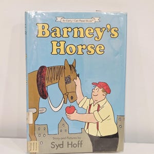Puede incluir: Un libro infantil titulado "Barney's Horse" con una cubierta azul. La ilustración de la portada muestra un caballo marrón y un hombre con gorra roja y tirantes. El hombre ofrece una manzana roja al caballo. El texto incluye "An Early I Can Read Book" y "Story and Pictures by Syd Hoff."