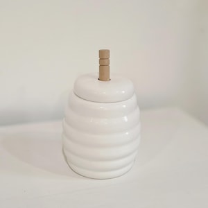May include: A white ceramic honey pot with a ridged body and a wooden handle on the lid. The pot has a glossy finish and a round shape. The wooden handle is cylindrical and made of stacked pieces.