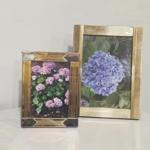 May include: Two rectangular picture frames with metallic gold-toned finishes. The smaller frame features silver-toned corner accents and contains a photograph of pink hydrangea flowers. The larger frame has a ribbed texture and holds a blue hydrangea.