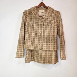 May include: A vintage two-piece suit in a tan and brown plaid pattern. The jacket has a rounded collar and three buttons. The skirt is a matching A-line style. The suit is hanging on a wooden hanger.