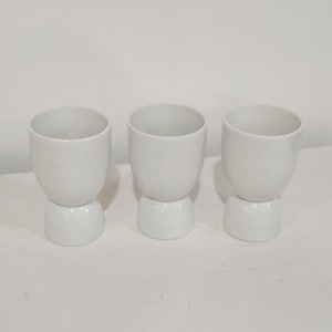 May include: Three white ceramic cups with a rounded top and a bulbous base are arranged on a white surface. The cups have a simple, modern design, and are likely used for drinking beverages.