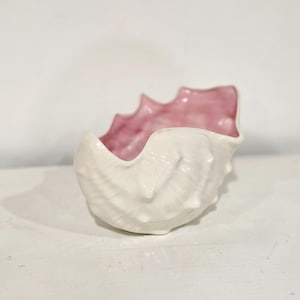 May include: A ceramic bowl shaped like a seashell with a textured, off-white exterior and a smooth, glossy light pink interior. The rim features a scalloped, irregular edge that mimics the natural form of a shell with raised ridges.