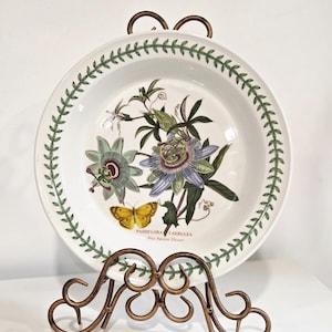 May include: A white decorative plate with a botanical design of passion flowers and a yellow butterfly, displayed on a bronze-colored metal stand. The plate has a green leaf border and text that reads "Passiflora Caerulea".