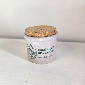 May include: A small, off-white ceramic jar with a cork lid. The jar is labeled "COLE SLAW SEASONING" with a net weight of 3.75 oz. A green and white circular logo is also visible on the jar.