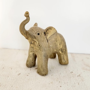 May include: A small, wooden elephant figurine with its trunk raised in the air. The elephant is painted in a light brown color and has a textured surface.