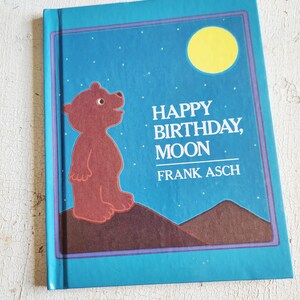 May include: A children's book titled "Happy Birthday, Moon" by Frank Asch. The cover features a brown bear looking up at a yellow moon against a blue starry sky. The book has a teal border.