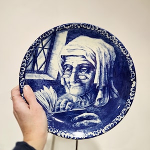 Puede incluir: Plato decorativo azul y blanco con una ilustración detallada de una persona mayor leyendo un libro. El plato tiene un borde festoneado con un diseño floral. La obra de arte recuerda a la cerámica de Delft.