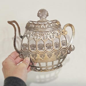 Op de afbeelding: Zilverkleurige decoratieve theepot met een ingewikkeld opengewerkt ontwerp. De theepot heeft een handvat, een tuit en een deksel met een schelpvormige bekroning. Het ontwerp bevat schelp- en bloemmotieven.