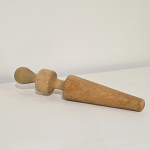 May include: A wooden rolling pin with a rounded handle and a tapered, cylindrical rolling surface. The rolling pin is made of light-colored wood and is lying on a white surface. The rolling pin is designed for use in baking.