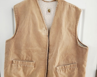 May include: A tan Carhartt vest with a white, fuzzy interior lining. The vest has two front pockets and a button closure. The Carhartt logo is visible on the interior lining. The vest is hanging on a gold-colored hanger.