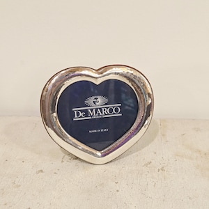 May include: A heart-shaped silver picture frame with a dark blue interior. The frame has a polished finish and features the text "De Marco Argenti" and "Made in Italy". The frame is designed to hold a photograph.