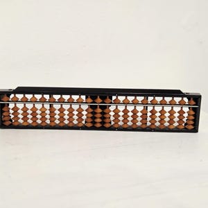 May include: A black abacus with wooden beads. The abacus is rectangular and has multiple rows of beads. The beads are a light brown color and are arranged in columns. The abacus is sitting on a white surface.