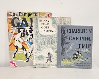 May include: Three vintage children's books titled The Camper's Game Book, Buzzy Bear Goes Camping, and Mr. Charlie's Camping Trip. The books feature illustrated covers with various camping scenes, including figures, a bear, and a campfire in blue, yellow, and white.