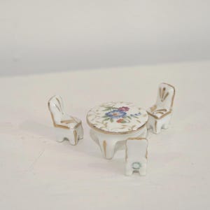 May include: Miniature porcelain tea set featuring a round table and three chairs. The table and chairs are white with gold trim and floral designs in blue, pink, and green. A charming addition to a dollhouse or miniature collection.