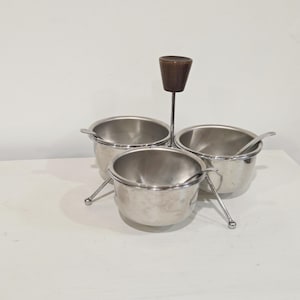 May include: A stainless steel condiment server featuring three round bowls arranged around a central metal rod topped with a dark brown wooden handle. Each bowl includes a small matching stainless steel spoon resting inside the container.
