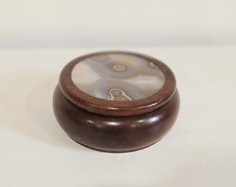 May include: A round, dark brown wooden box with a smooth, polished finish. The lid features a circular, inlaid stone disc with concentric, swirling patterns in shades of gray, white, and tan, creating a natural, banded appearance on the surface.