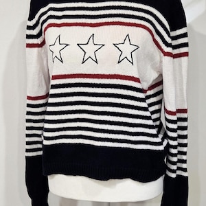 Könnte beinhalten: Ein langärmeliger Strickpullover mit einem horizontalen Streifenmuster in Marineblau, Weiß und dünnen roten Linien. Im Brustbereich befinden sich drei gestickte schwarze Sternumrisse. Der Pullover hat einen Rundhalsausschnitt mit rotem Rand.