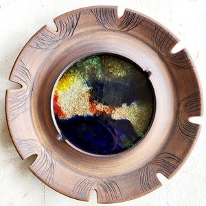 May include: A brown ceramic ashtray with a circular design. The center features a colorful abstract design with blue, yellow, red, and green hues. The outer rim has carved details and notched edges.