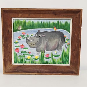 May include: A framed painting featuring a gray hippopotamus standing in a pond with pink and yellow water lilies. A small bird sits on the hippo's back, and a frog is visible on the grassy bank. The artwork is enclosed in a rustic brown wooden frame.