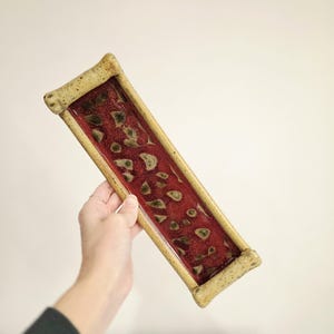 May include: A rectangular ceramic tray with a deep red interior speckled with green and tan shapes. The tray has a tan border and rounded corners. The tray is held against a white background.