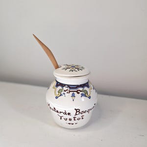 May include: A white ceramic mustard pot with a lid and a wooden spoon. The pot is decorated with blue, yellow, and green floral patterns and the words "Moutarde Bocquet, Yvetot".