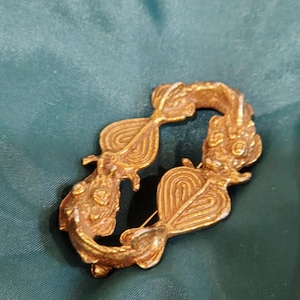 May include: A vintage gold-toned brooch with an ornate, openwork design. The brooch features a stylized, symmetrical shape with swirling patterns and floral accents. The pin is set against a dark green fabric background.