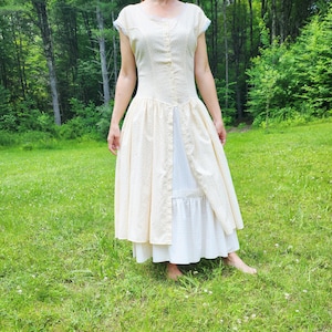 May include: A vintage-style dress in a cream color with a delicate floral pattern. The dress features short sleeves with lace trim, a button-down bodice, and a flowing skirt. The skirt has a white underskirt with a ruffled hem, creating a layered effect. The dress is worn outdoors.