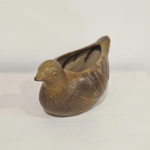 Könnte beinhalten: Ein brauner, vogelgeformter Keramikübertopf mit einer texturierten, gesprenkelten Glasuroberfläche. Das Gefäß verfügt über einen ausgehöhlten Rücken zur Aufnahme kleiner Gegenstände oder Pflanzen sowie geformte Flügeldetails und einen Kopf.