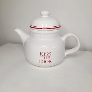 May include: White ceramic teapot with a lid and handle. The teapot has a red stripe around the lid and the words "KISS THE COOK" in red lettering on the front. The teapot is on a white surface.