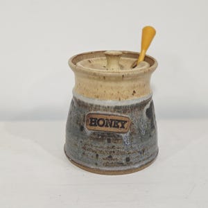 May include: A ceramic honey pot with a speckled beige lid and a dark grey, blue, and brown mottled base. The word HONEY is embossed on a rectangular plaque on the front. A small yellow wooden honey dipper rests inside the pot through the lid opening.