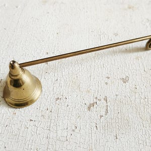 Peut inclure: Un éteignoir à bougie en laiton vintage. L'accessoire de bougie a une base en forme de cloche et une longue poignée se terminant par une spirale. Il est en laiton et repose sur une surface blanche vieillie.