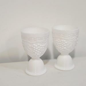 May include: Two white, vintage-style goblets. The goblets have a detailed grape and vine pattern, with a scalloped design at the rim. The stem is a rounded, pedestal base. The goblets are made of opaque white glass.