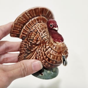 Pode incluir: Uma estatueta de peru em cerâmica com acabamento castanho brilhante, detalhes de penas texturizadas, cabeça e carúncula vermelho-escuras e um pequeno bico preto e branco. O peru assenta numa base arredondada verde mosqueada e lisa.
