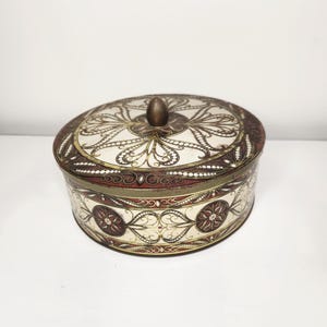 May include: An oval-shaped vintage tin box with a lid. The box features an ornate design in cream, brown, and gold. The lid has a central knob. The sides of the box are decorated with floral patterns and geometric shapes.